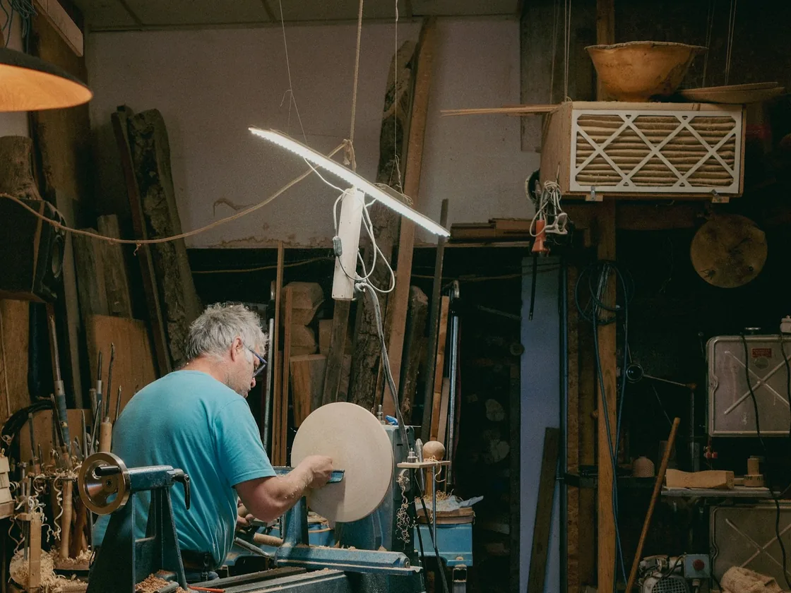 Taller de madera con torno, tablas apiladas y un disco de madera clara sujeto entre puntos